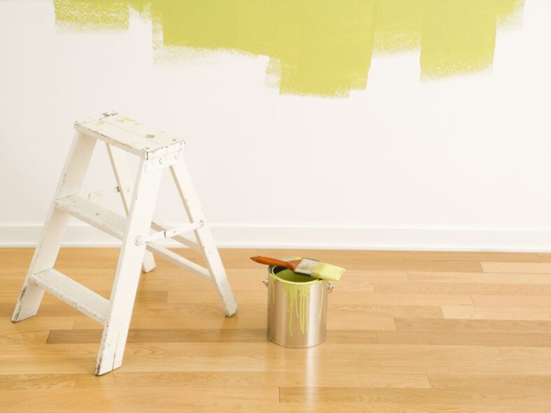 a foot stool, bucket of paint, and brush in front of a wall with yellow paint